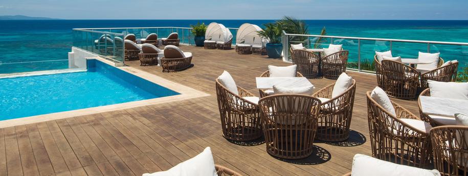 בריכת אינפיניטי במרפסת המלון עם דק עץ מסביב וכסאות ישיבה עץ וכריות לבנות מול נוף הים