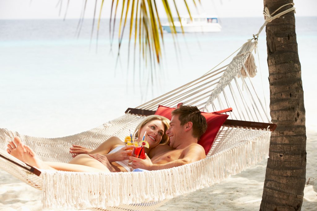 זוג שוכב על ערסל עם משקאות ביד על חוף אקזוטי