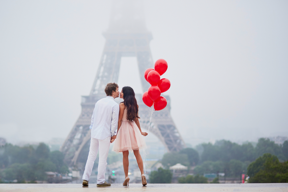 זוג מביא נשיקה, הבחורה מחזיקה בלוני הליום אדומים ובנוף ערפל ורואים קצת את מגדל אייפל