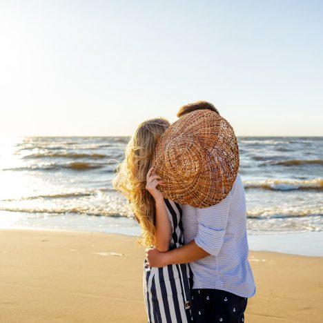 זוג עומד מחובק מול הים ומכסה את הפניה מאחורי כובע