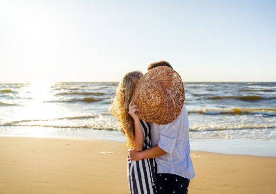 זוג עומד מחובק מול הים ומכסה את הפניה מאחורי כובע